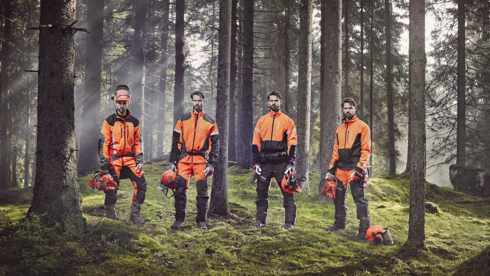 4 men standing in a forest, all wearing full garden PPE, including helmet, boots and chainsaw clothing