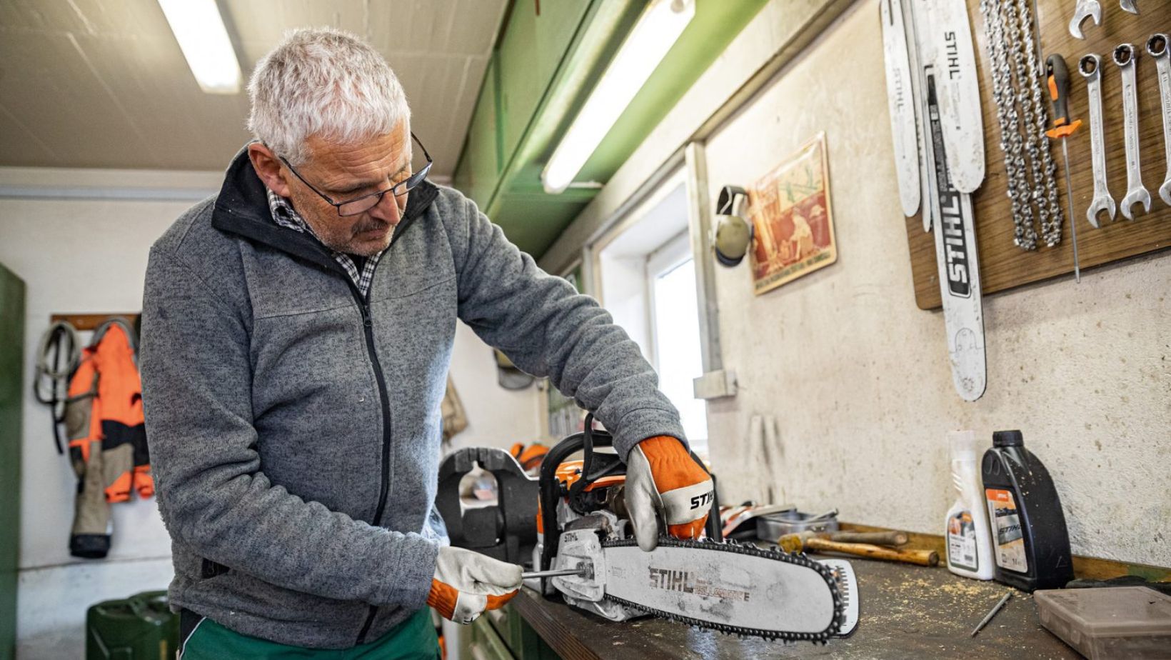 a man with short grey hair and glasses, stood in a workshop, tensioning a chainsaw chain