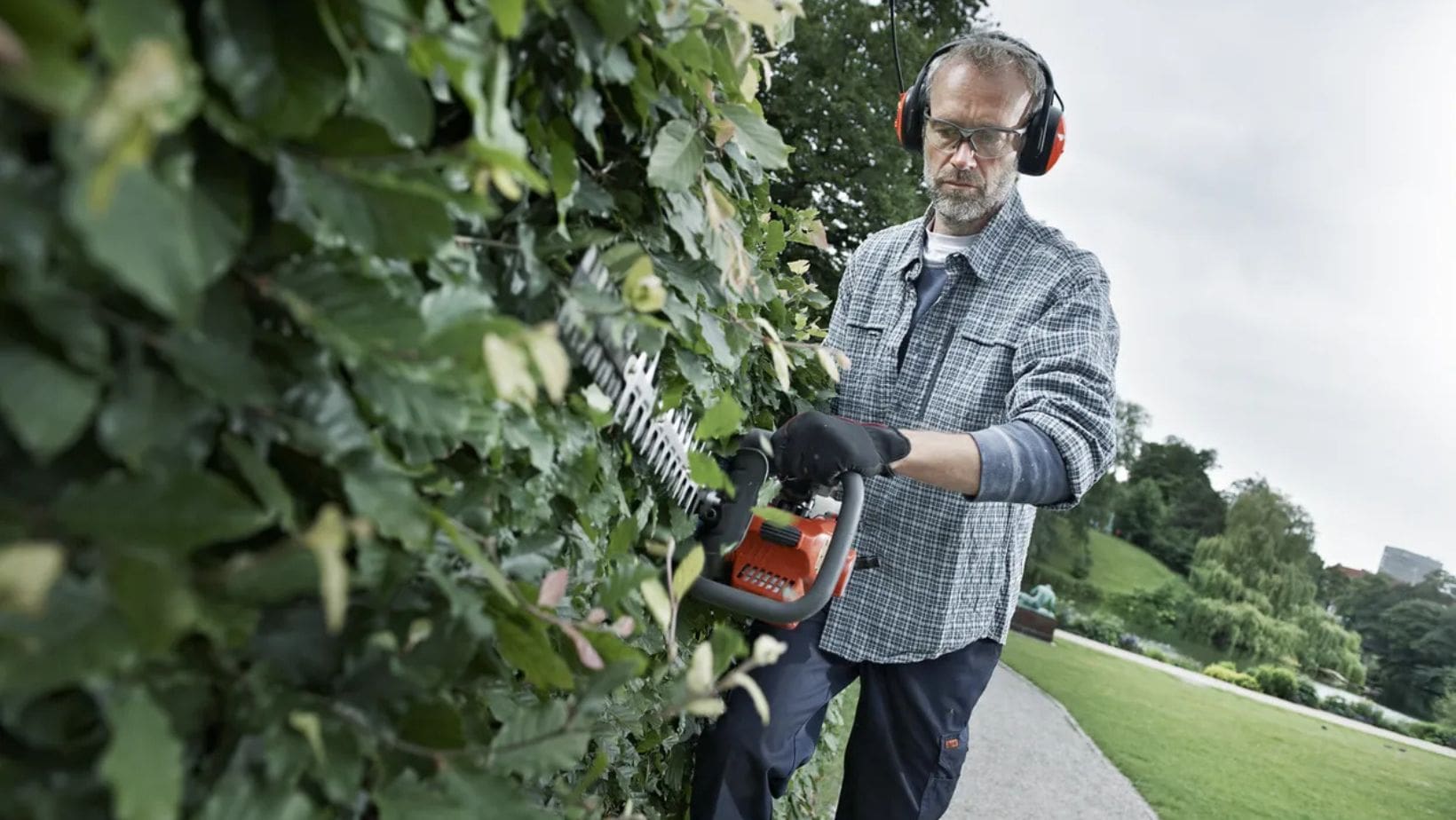 a man wearing ear defenders, safety glasses and gloves, using a hedge trimmer