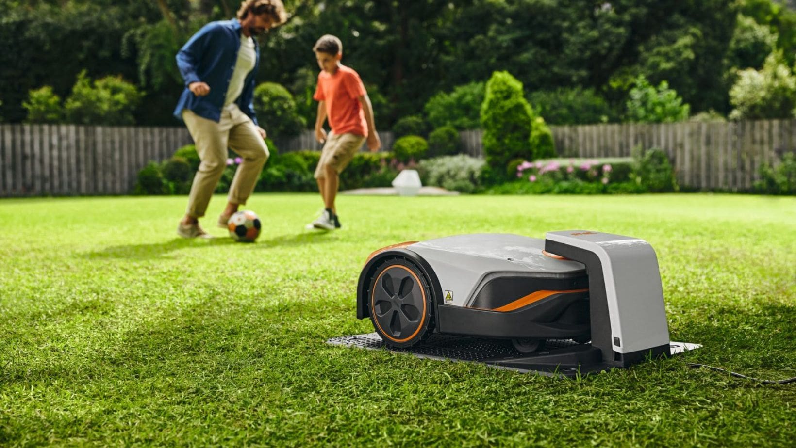 a STIHL iMow robotic lawn mower in its charging station on a lawn, with a father and son playing football in the background